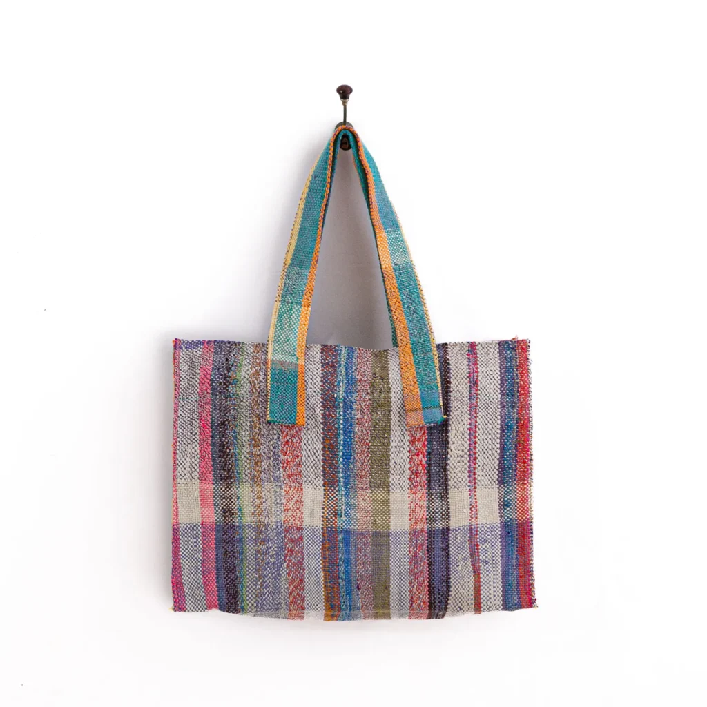 A multicolored woven tote bag with vertical stripes of various patterns and shades hangs on a wall hook.