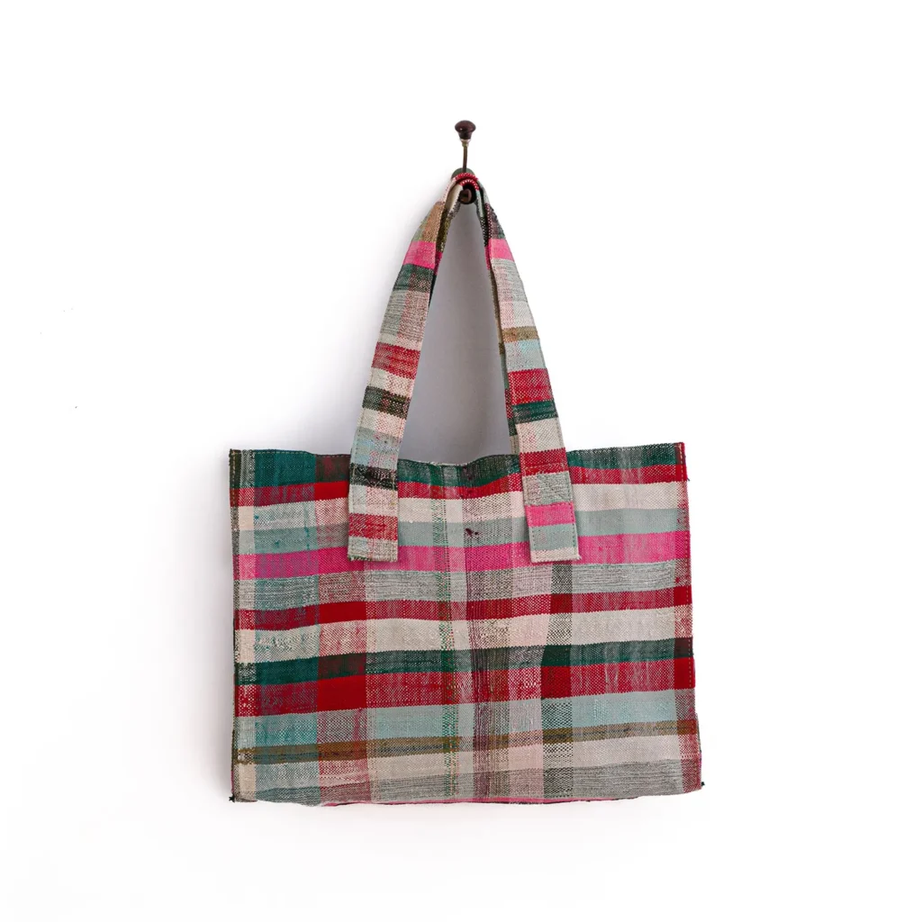 A multicolored plaid tote bag hangs on a hook, featuring red, pink, green, and gray stripes.