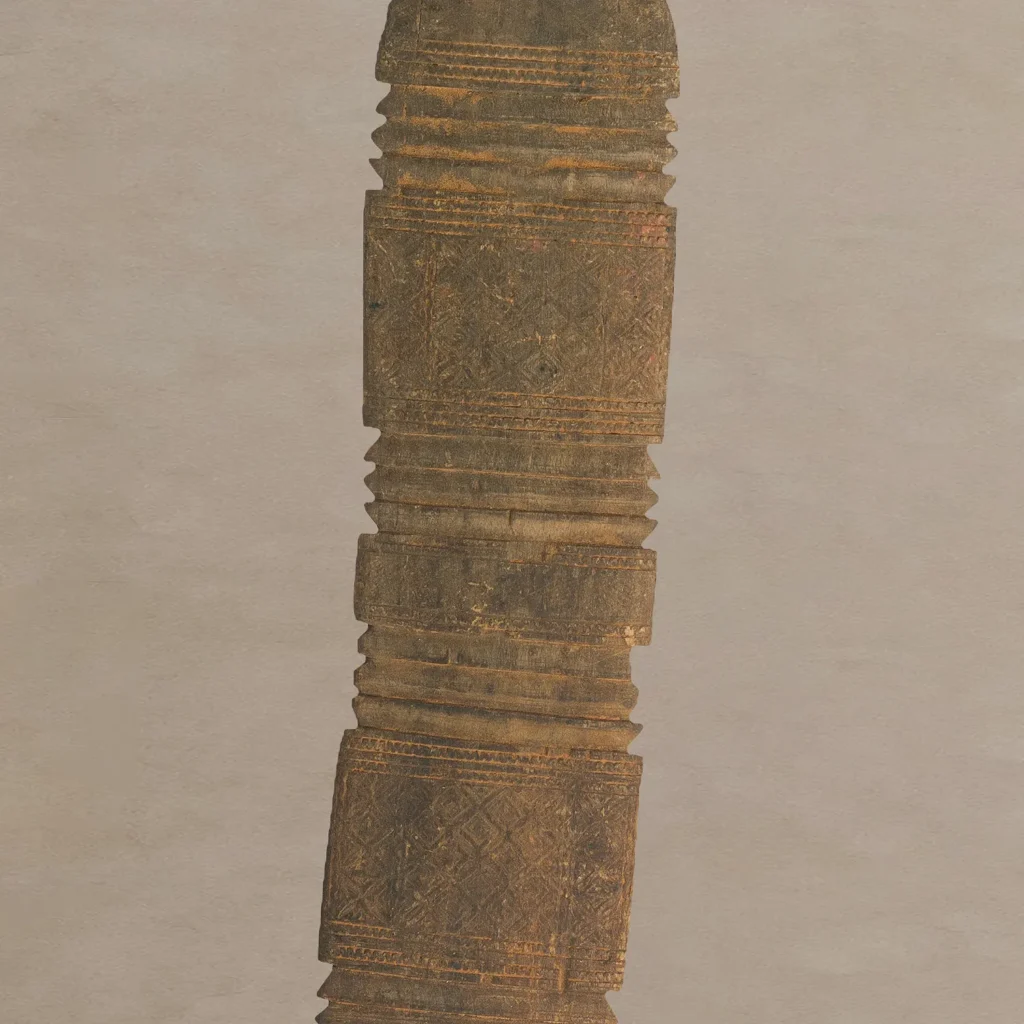 Close-up view of a hand-carved wooden column with intricate patterns and vertical grooves reminiscent of a vintage tent peg against a plain background.