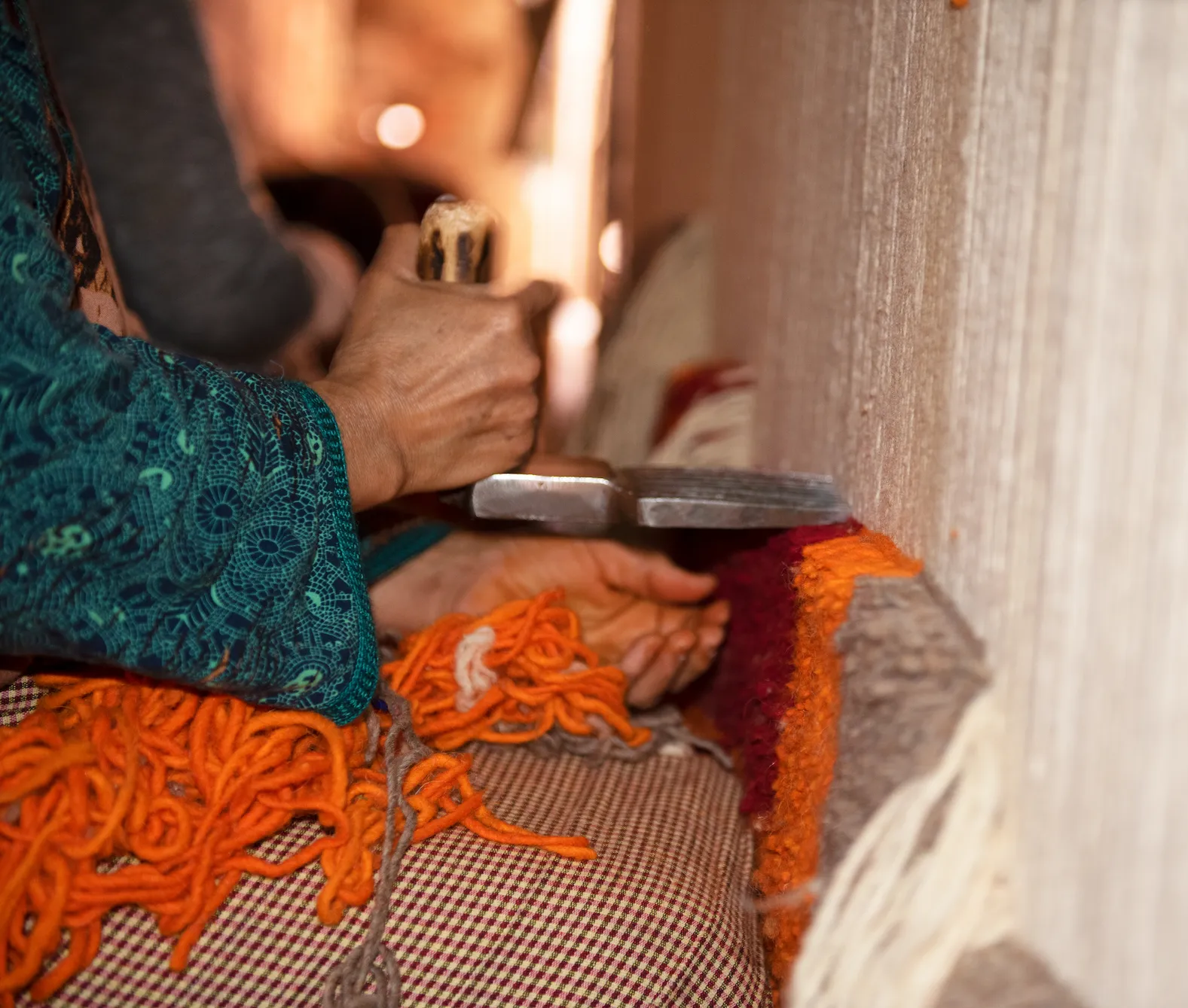 Close-up of a person weaving a carpet by hand, pressing threads with a metal tool on a loom. Orange and red yarn are visible.