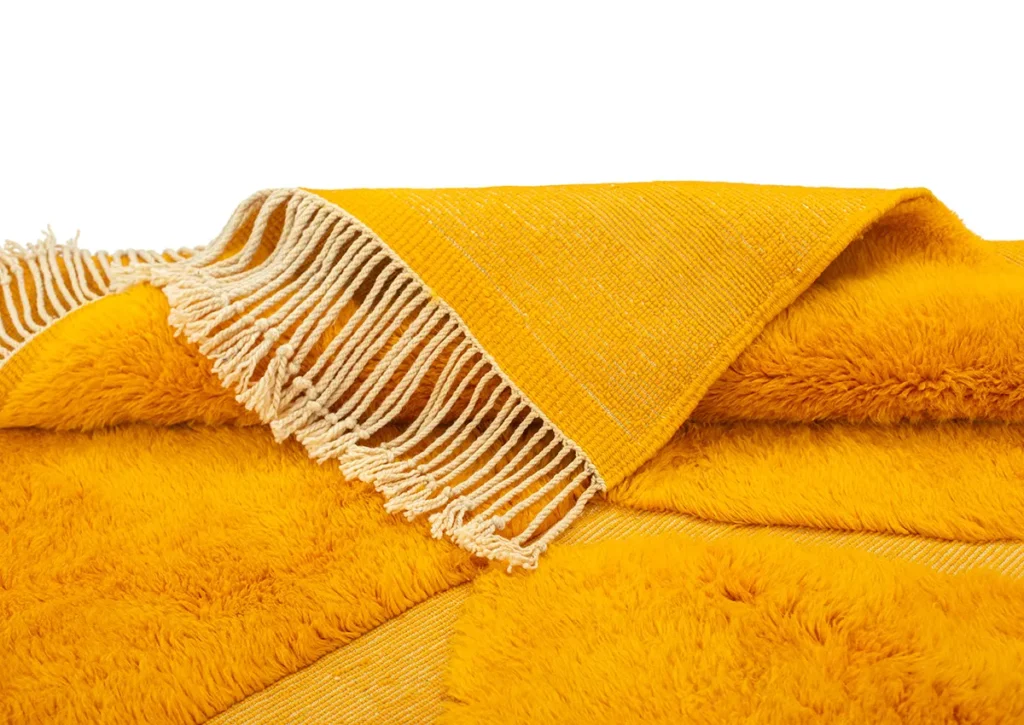 A hand-knotted rug in a warm yellow hue, with a fringe edge and featuring a mix of smooth fabric and plush, furry texture, offering a touch of Moroccan luxury to your space.