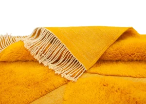 A hand-knotted rug in a warm yellow hue, with a fringe edge and featuring a mix of smooth fabric and plush, furry texture, offering a touch of Moroccan luxury to your space.