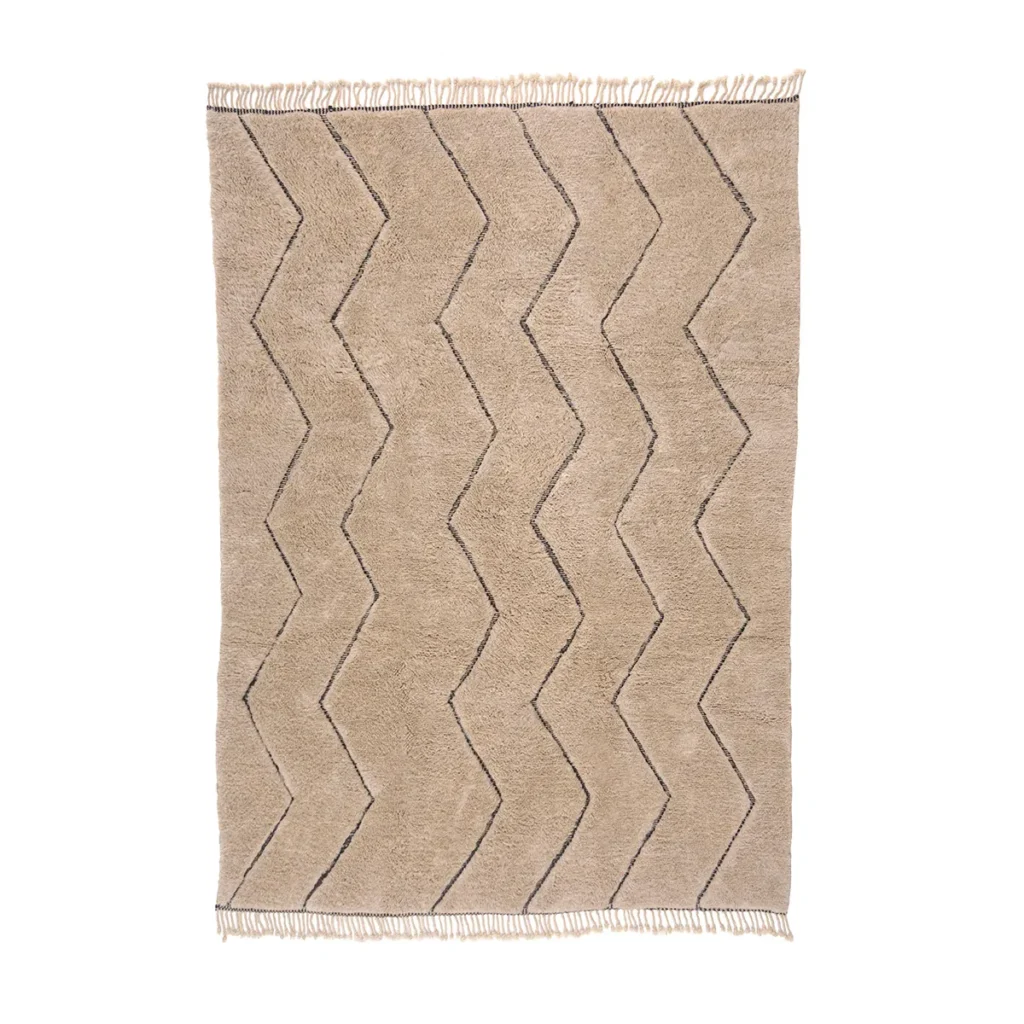 This authentic Moroccan rug is hand-knotted with a beige textured base, featuring a zigzag pattern in dark lines. The edges are adorned with fringe detailing, and its design showcases alternating chevrons for an artisanal touch.