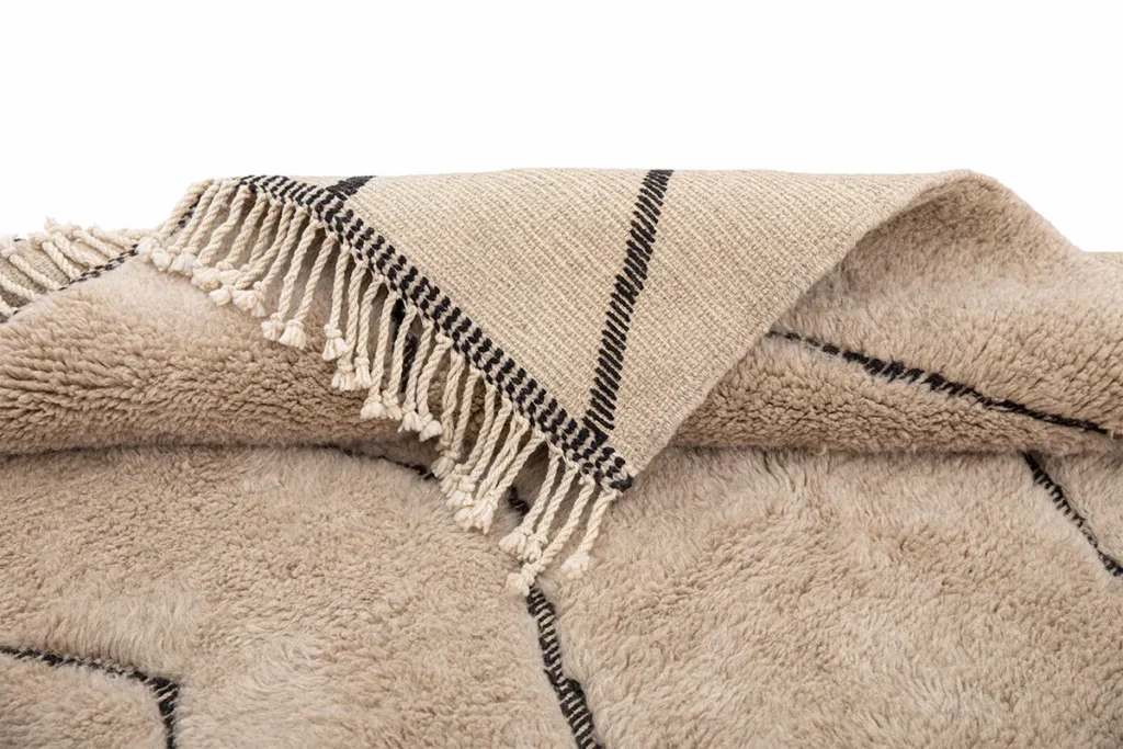 A hand-knotted textile with black lines and fringed edges is partially folded over a fuzzy, cream-colored fabric featuring a geometric pattern.