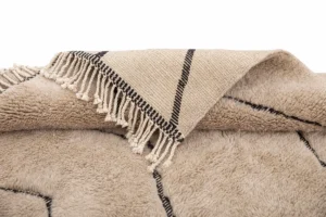 A hand-knotted textile with black lines and fringed edges is partially folded over a fuzzy, cream-colored fabric featuring a geometric pattern.