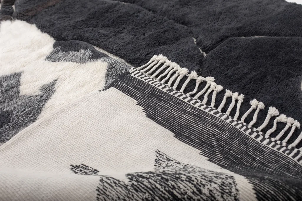 Close-up of a hand-knotted black and white woven fabric with a fringed edge and geometric patterns. This artisanal rug showcases the intricate craftsmanship typical of authentic Moroccan rugs.