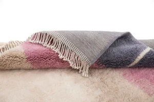 A luxurious, hand-knotted rug with fringe, showcasing shades of pink, beige, gray, and blue, lies elegantly on a fluffy cream-colored carpet.