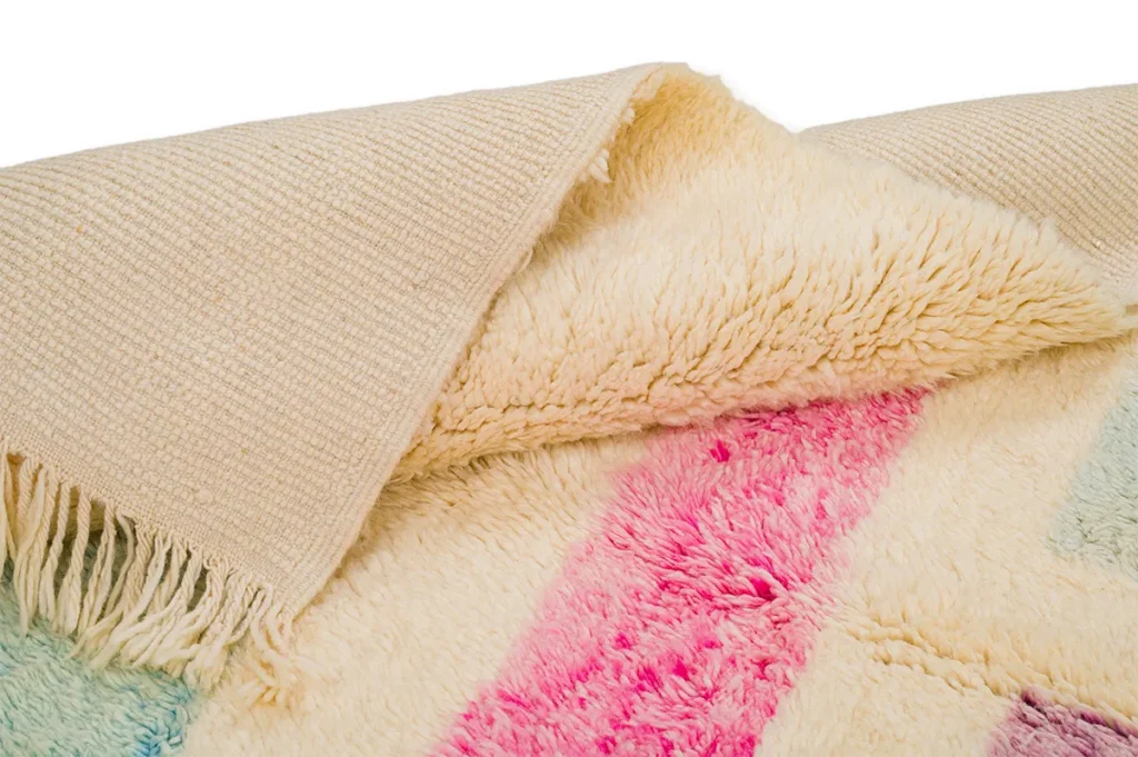 Close-up of a textured woven beige fabric partially covering a fluffy cream rug with pink and blue stripes. Fringed edges are visible, adding an artistic touch.