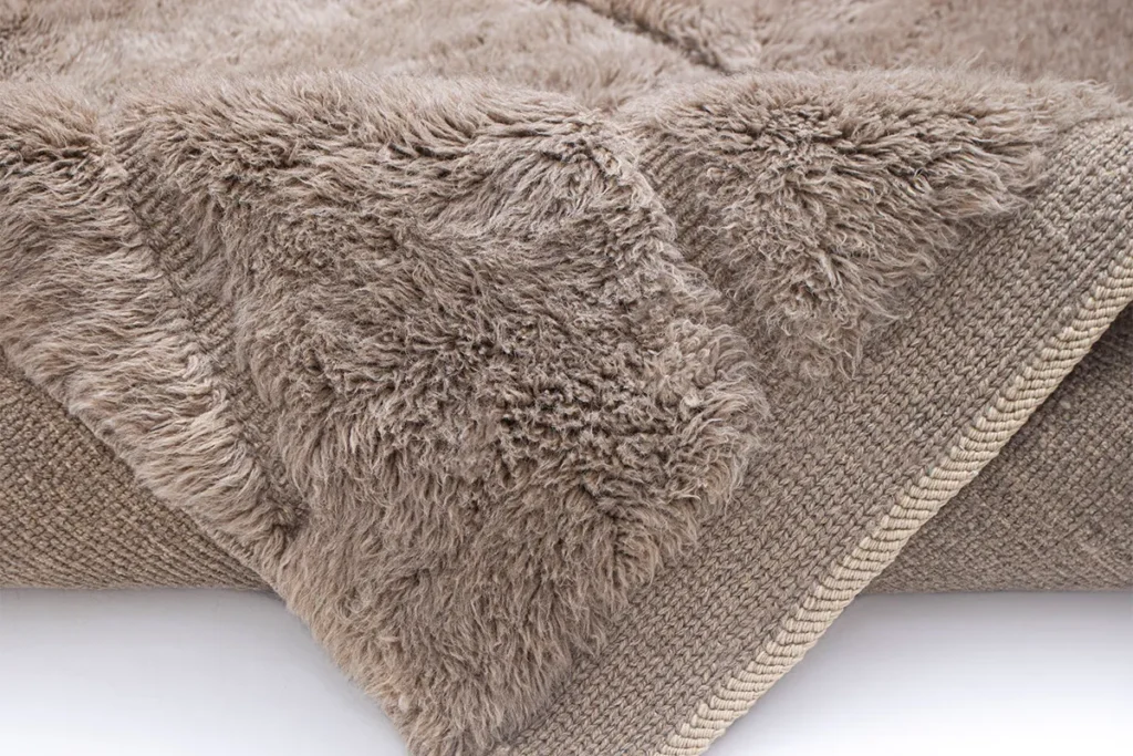 Close-up of a beige, fluffy, textured piece with a woven underside, folded at the edge. This luxurious fabric captures the essence of a hand-knotted Moroccan rug, offering both style and comfort to any space.