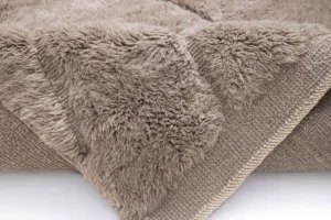 Close-up of a beige, fluffy, textured piece with a woven underside, folded at the edge. This luxurious fabric captures the essence of a hand-knotted Moroccan rug, offering both style and comfort to any space.