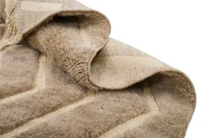 Close-up of a folded, beige textured luxury rug with a chevron pattern. It has a soft, plush surface and a woven underside, epitomizing elegance and comfort.