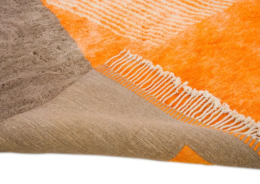 A close-up of a luxurious, hand-knotted rug showcases its vibrant orange, gray, and beige textures. One corner is flipped over to reveal the intricate underside. Long white fringes elegantly line one edge.