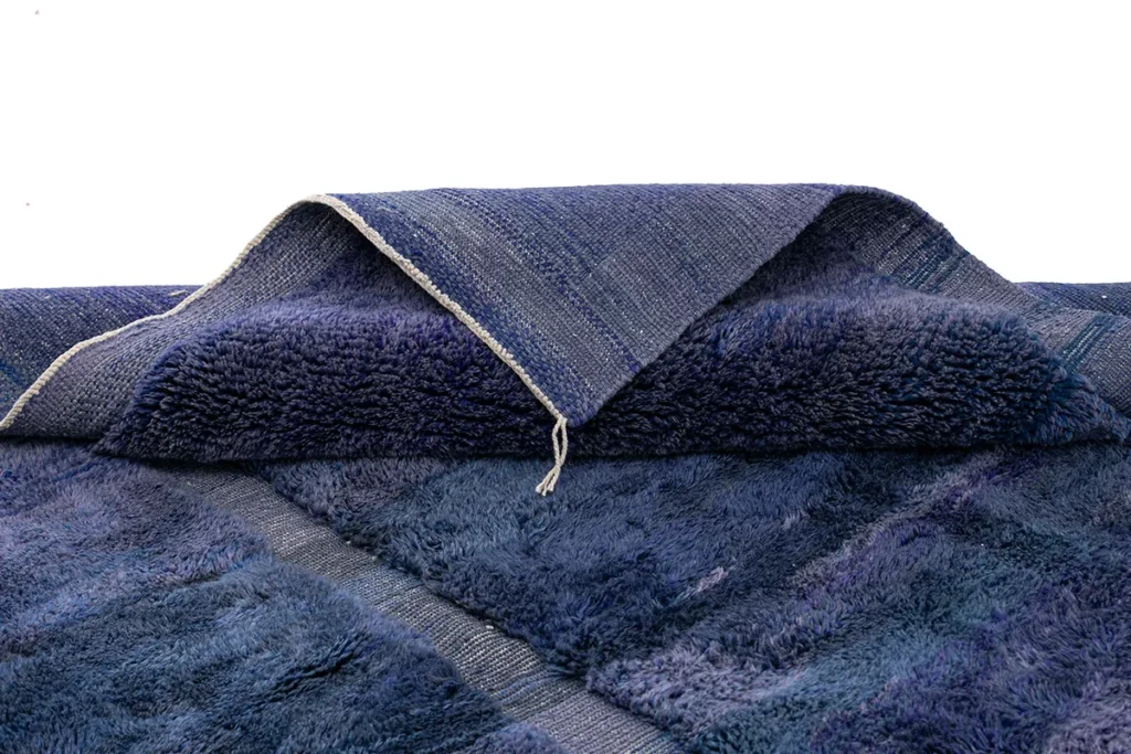 This luxury rug boasts a folded blue shaggy surface with striking geometric patterns, revealing a lighter fabric underside. Its elegant design and premium quality make it an exceptional addition to any interior, set against a crisp white background.