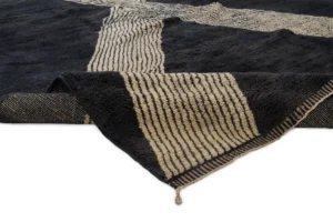 A close-up of an authentic Moroccan rug featuring a black and cream striped texture. The corner is folded, revealing the artisanal underside pattern that highlights its hand-knotted craftsmanship.