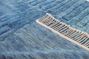 This luxury rug features a close-up of a blue textured surface with a striped pattern. One edge boasts beige fringe, adding a decorative element to the hand-knotted design.