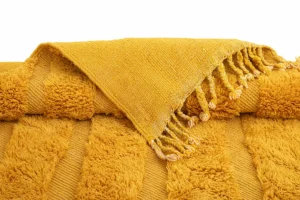 A textured yellow piece with fringe edges is stacked elegantly over another, both exuding an aura of opulence reminiscent of luxury rugs.