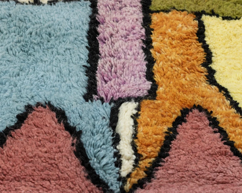 Close-up of a colorful, hand-knotted rug with abstract shapes in blue, purple, orange, pink, and green, outlined in black. This artisanal piece showcases the intricate craftsmanship found in authentic Moroccan rugs.
