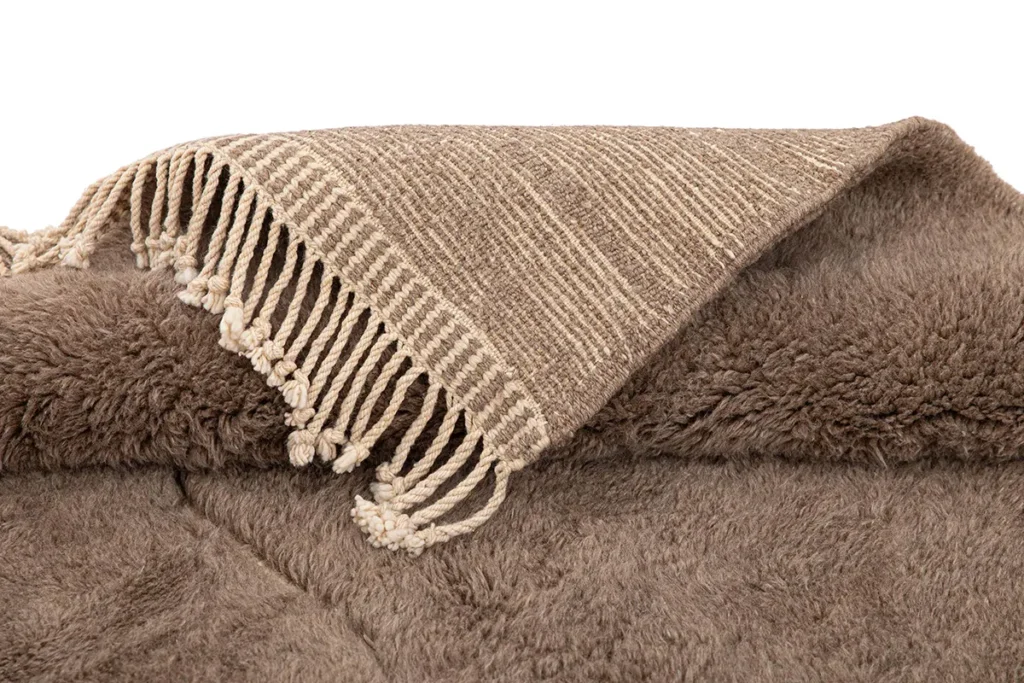 A hand-knotted Moroccan rug in shades of brown and beige lies partially folded, showcasing its textured fabric and fringe edges.