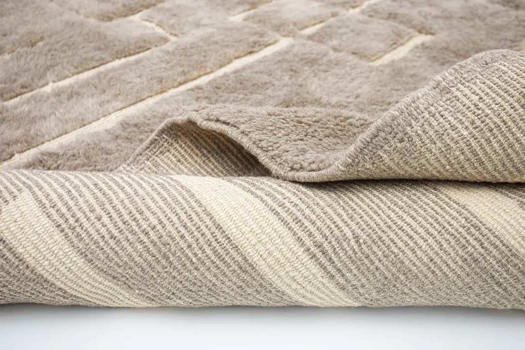 This close-up showcases a hand-knotted, beige and cream luxury rug, featuring a textured surface and striped underside, elegantly placed on a smooth white background.