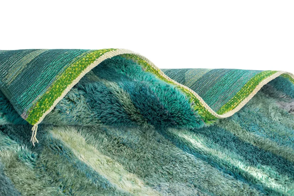 This luxurious hand-knotted rug features a close-up view of its textured, fuzzy surface in vibrant green and blue hues. The distinctive striped pattern and slightly raised edges reveal its expertly woven underside, adding an elegant touch to any room.