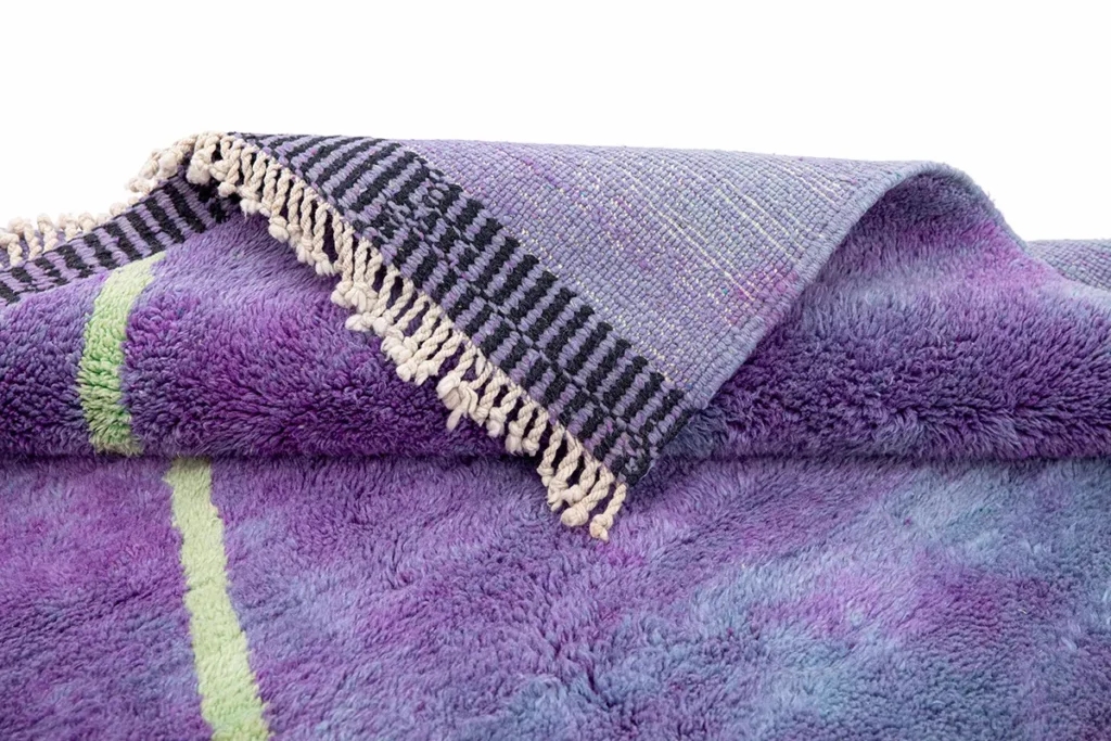 A close-up of a luxurious, hand-knotted purple rug with fringed edges and black stripes displays various shades and a green line.