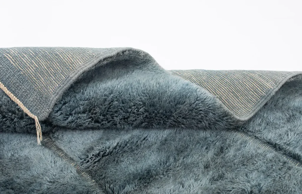 A close-up of an artisanal, teal shag rug folded over reveals both its fuzzy pile and the intricate woven underside, showcasing hand-knotted craftsmanship.