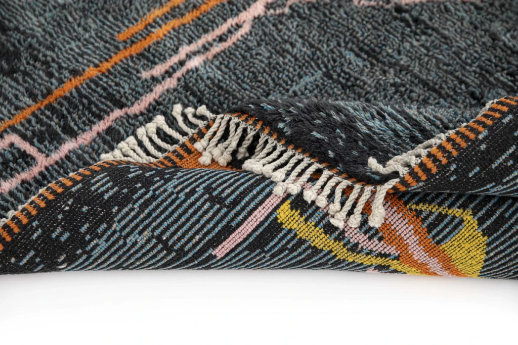 Close-up of a luxury hand-knotted rug with a corner elegantly folded over. The design showcases abstract patterns in orange, pink, yellow, and white against a dark background. White fringes gracefully accent the edge.