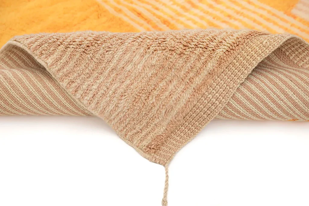Two folded layers rest on each other; the top is a light brown piece with a string loop, and the bottom boasts stripes of orange and white. This elegant setup evokes the feel of luxury rugs, blending subtle design with refined sophistication.
