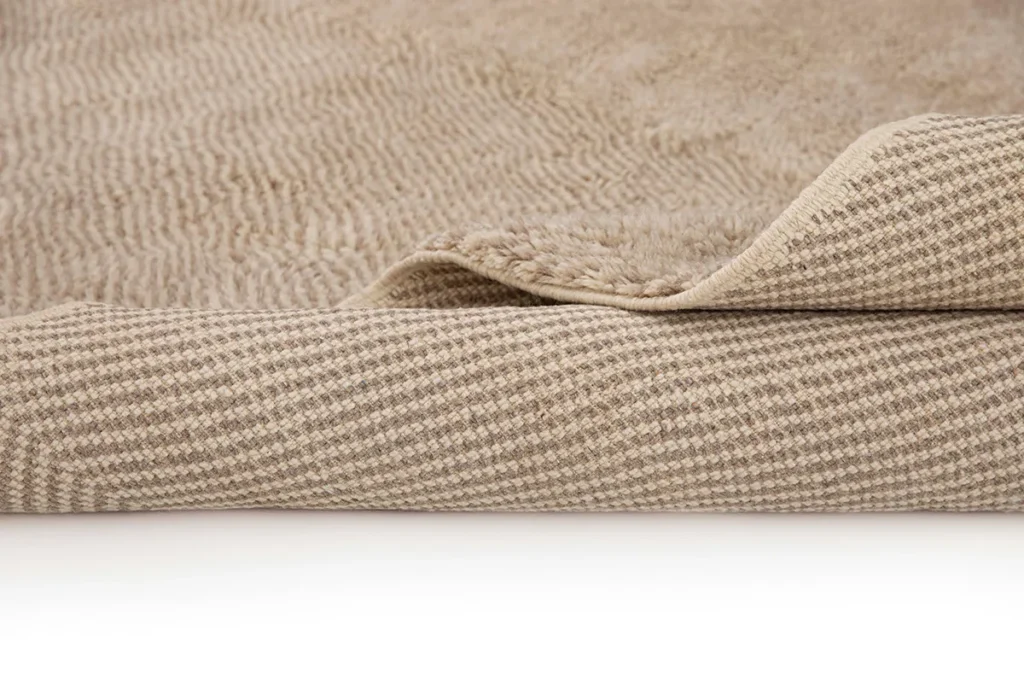 This luxurious rug features a close-up of its beige textured surface with a folded corner, revealing an intricate weave pattern underneath. The top is exceptionally soft, embodying the elegance of a hand-knotted Moroccan design.