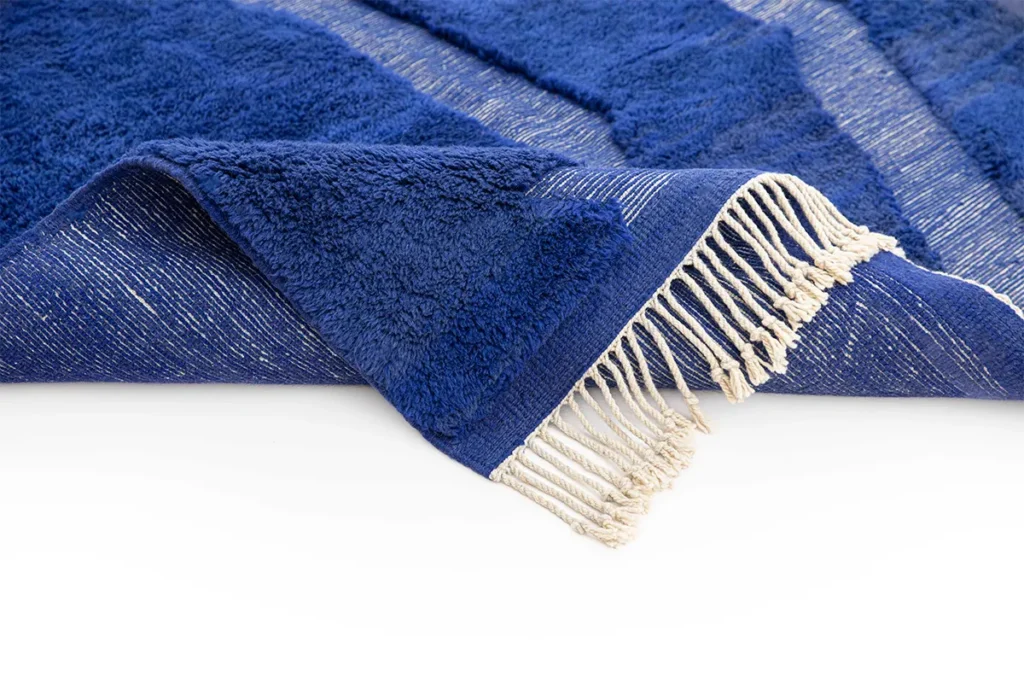Close-up of a blue plush fabric with fringe detailing on one edge, layered over another similar piece, displayed against a plain white background. The texture exudes the luxurious quality reminiscent of a hand-knotted Moroccan rug.