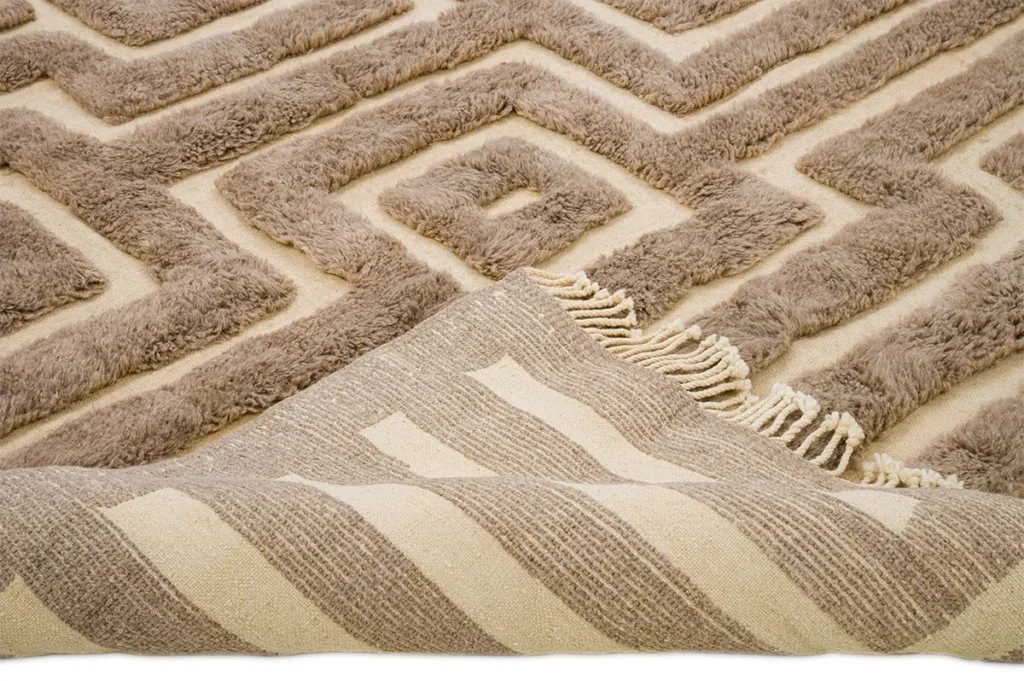 A close-up of a hand-knotted, textured beige rug showcases its geometric pattern. The corner is folded back to reveal the underside with a striped design and fringe, enhancing the allure of this luxury Moroccan rug.