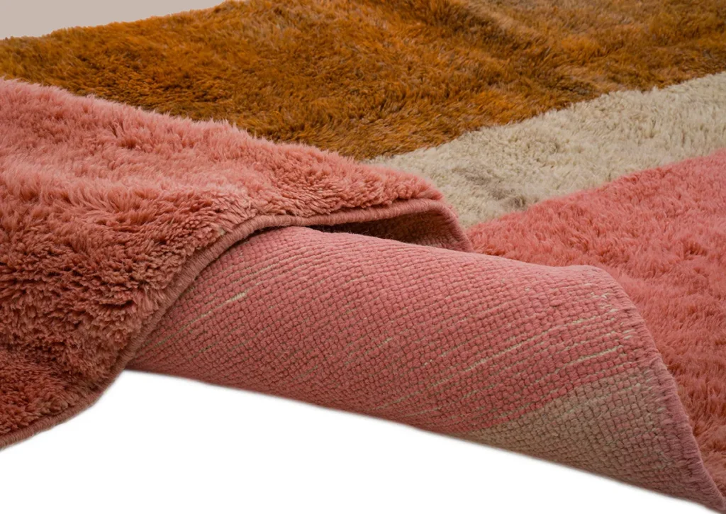 A close-up of a folded, multicolored luxury rug reveals its vibrant textures. This exquisite piece, featuring pink, brown, and beige hues, showcases both the plush pile and the intricate hand-knotted underside.