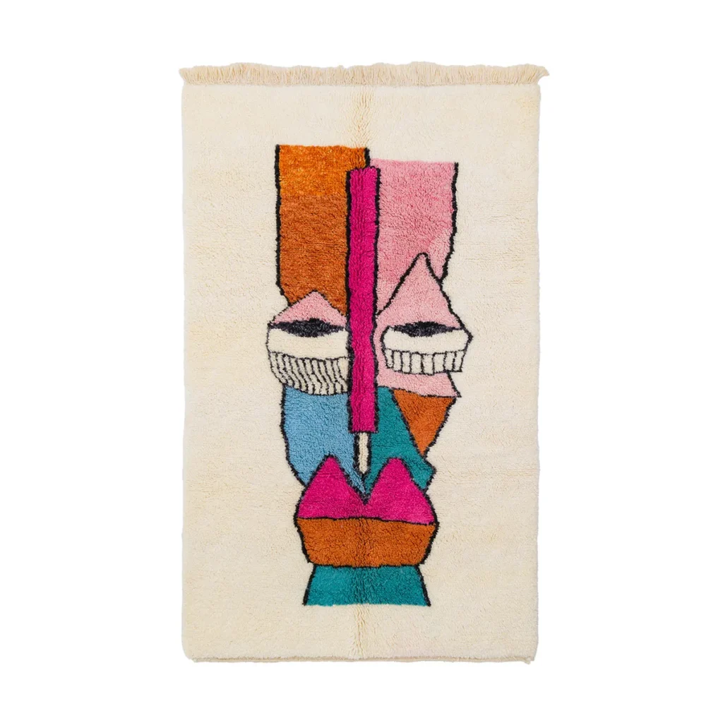 This luxury rug features an abstract, colorful face design in pink, orange, blue, and black on a cream background.
