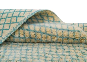 Close-up of a folded, beige and teal checkered rug. The top displays a flat pattern, while the underside is luxuriously fluffy, maintaining the same design. Perfect for adding style and comfort to any room.