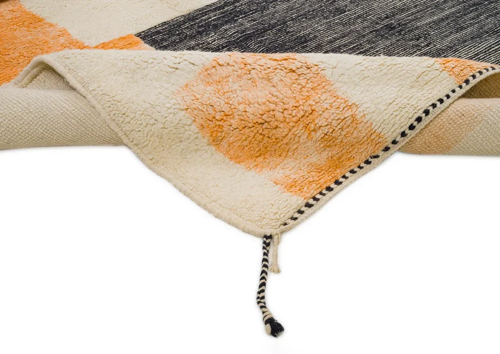 A close-up of a folded, textured rug reveals an orange circular pattern with black striped edges. A black and white braided string elegantly accents the corner, highlighting the intricate craftsmanship and vibrant design.