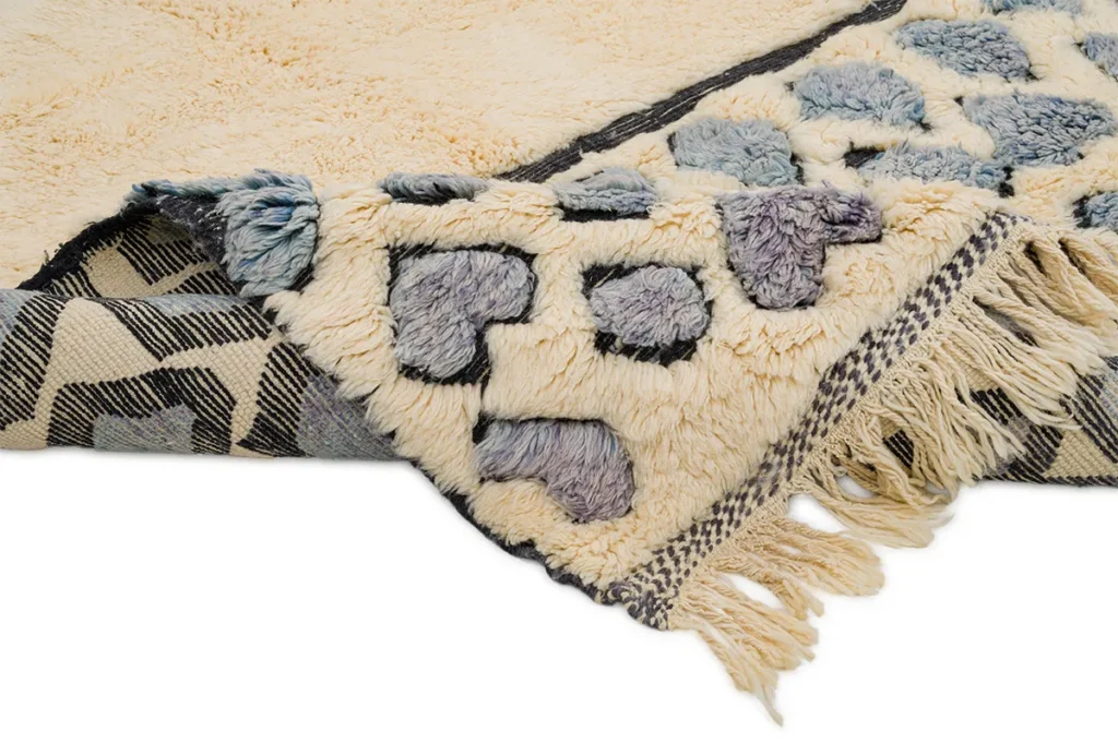 A close-up of a hand-knotted rug reveals a fringed corner, showcasing its intricate geometric pattern in cream, blue, and gray tones.