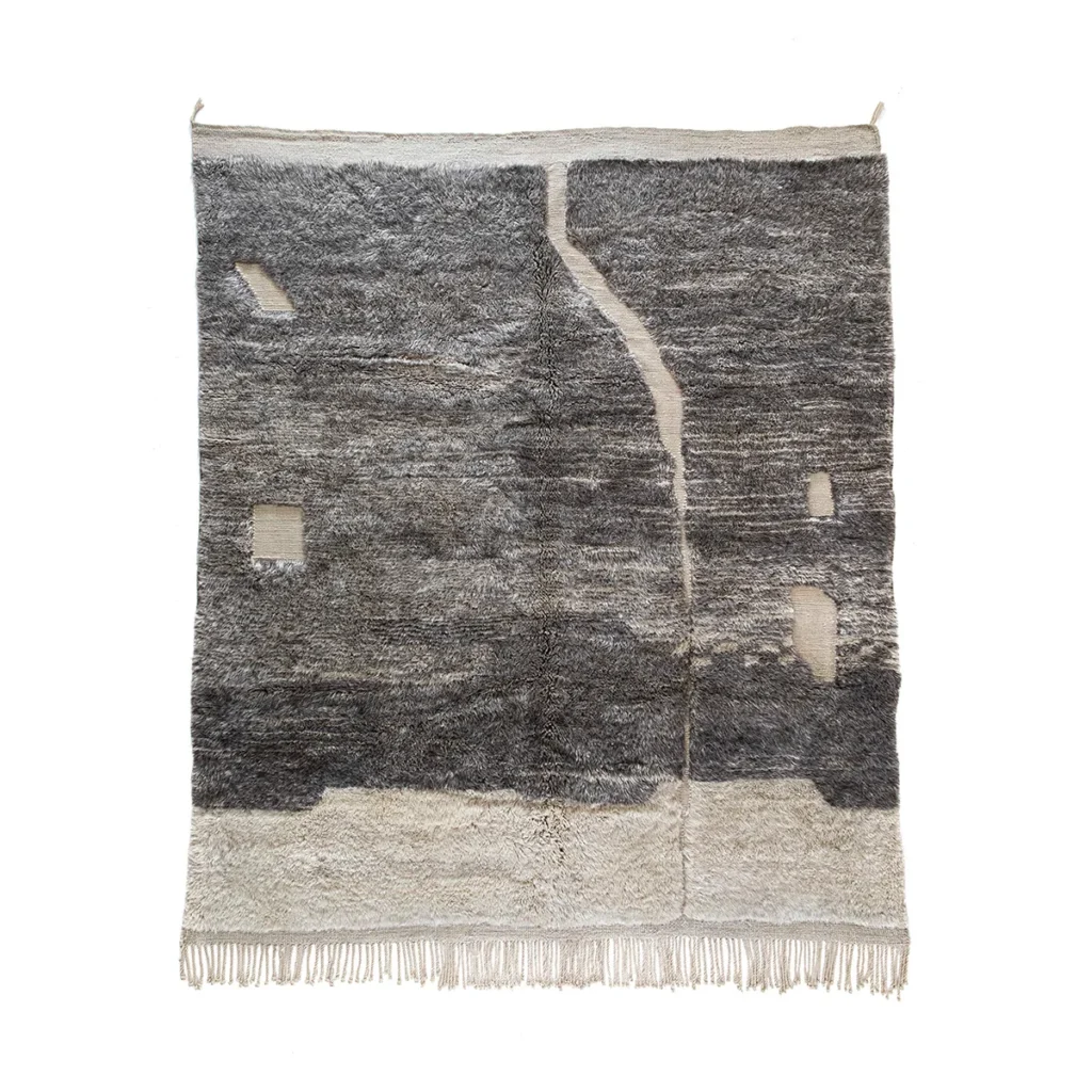 A textured, woven artisanal rug with a blend of gray and beige tones, featuring abstract patterns and fringed edges at the bottom.