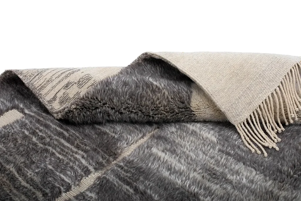 This authentic Moroccan rug features a folded, patterned gray and cream design with fringe detail. The texture is fluffy with a woven underside, showcasing its handcrafted artistry.