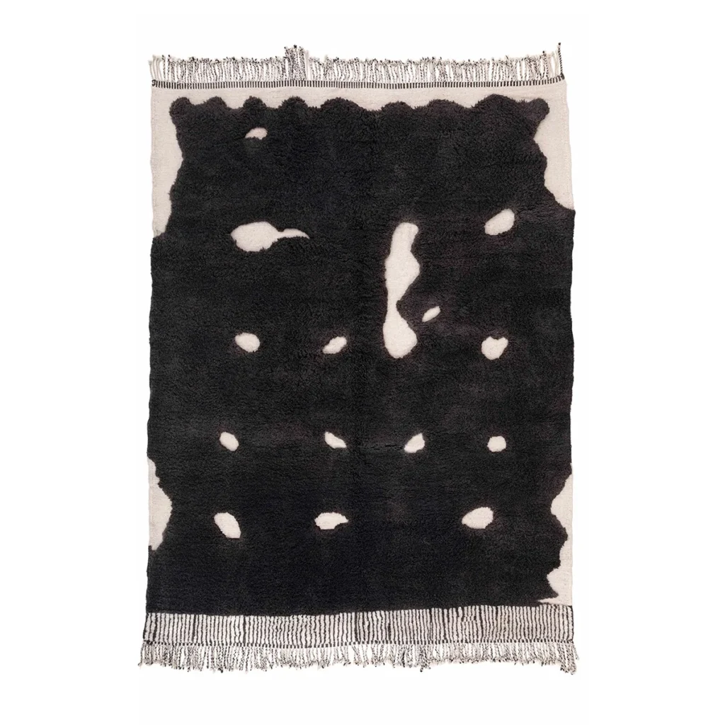 An artisanal rectangular rug with a black abstract pattern featuring irregular white spots, framed by fringed edges, reminiscent of authentic Moroccan rugs.