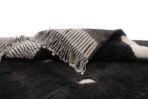 A close-up of an artisanal rug with a black and white checkered pattern and fringed edges, resting on a fluffy black surface.
