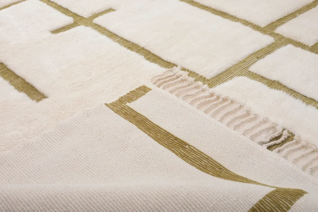 Close-up of an artisanal rug with green geometric lines, featuring fringed edges. The corner is folded to reveal its hand-knotted underside with a matching pattern, reminiscent of authentic Moroccan rugs.