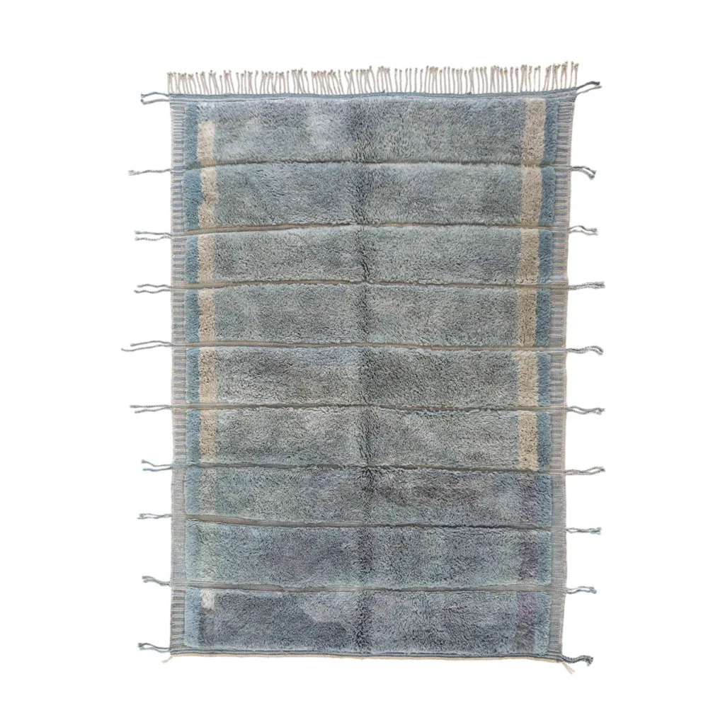 A rectangular, blue-gray rug featuring a textured surface with fringe on both ends and lighter vertical stripes along the edges. The elegant design of this decorative piece adds a touch of sophistication to any space.