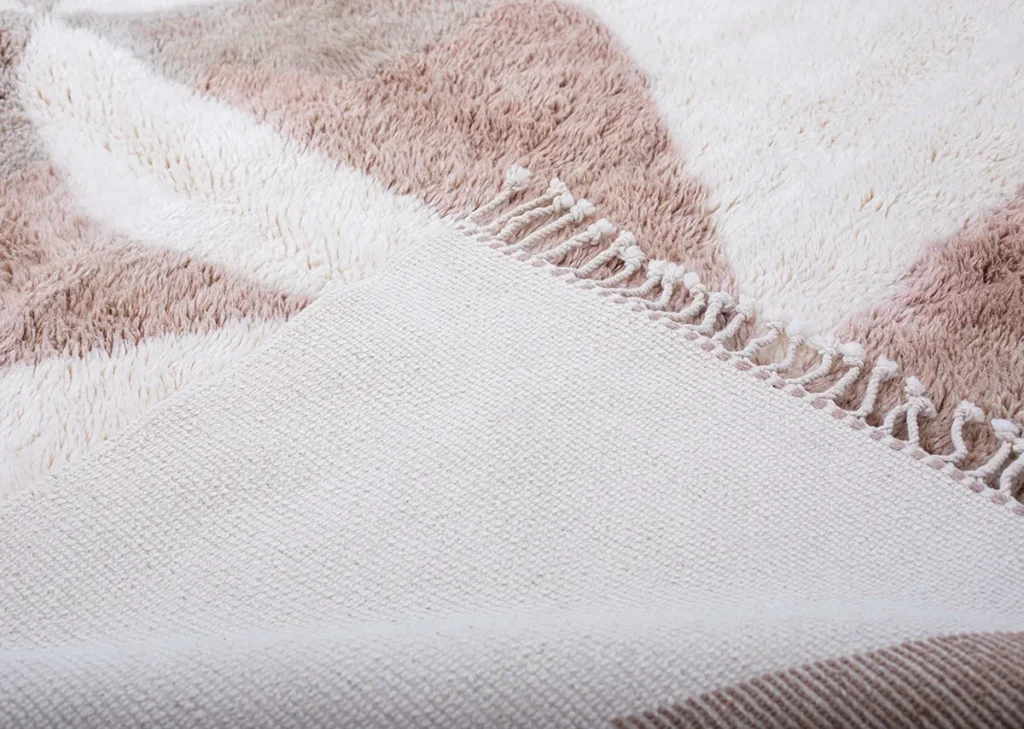 Close-up of a hand-knotted Moroccan rug showcasing a soft, patterned textile in beige, cream, and light brown tones. The edge features short fringed tassels, adding an elegant touch.