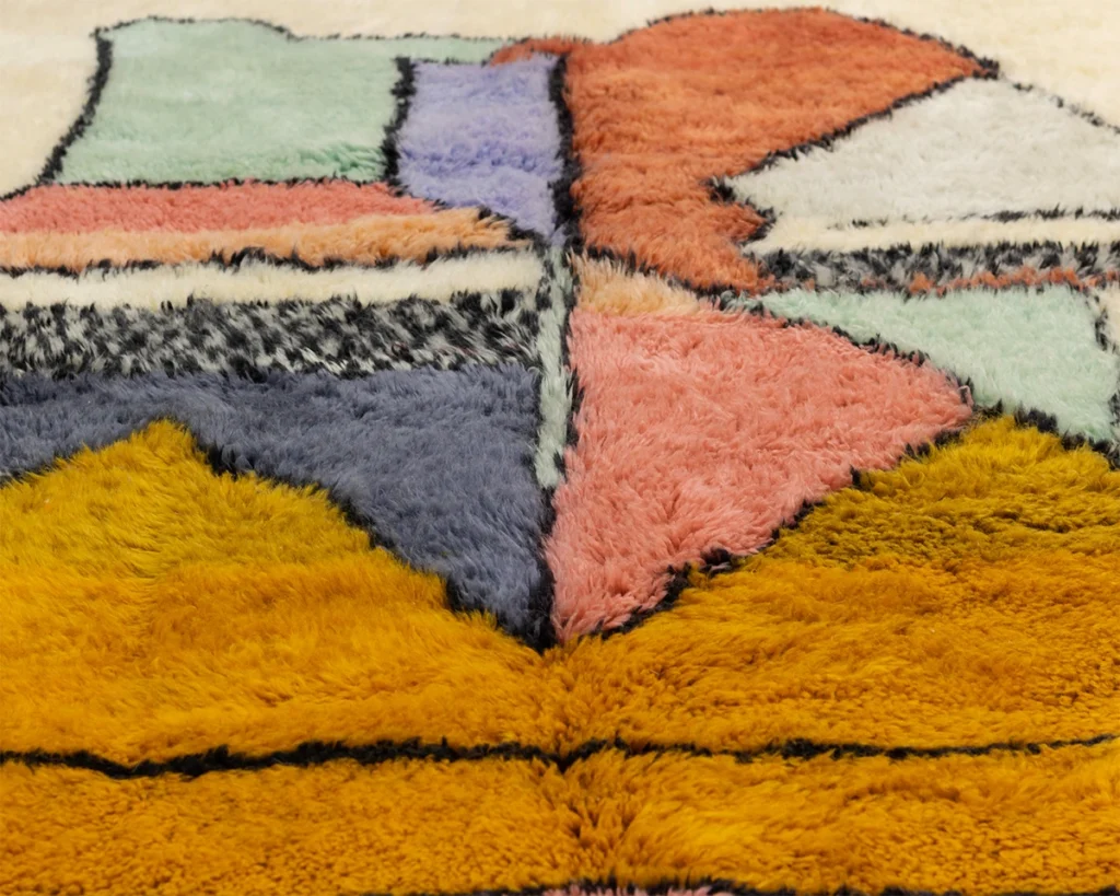 This close-up reveals a hand-knotted, colorful rug with a geometric pattern featuring vibrant sections of orange, pink, blue, purple, and cream outlined in black.
