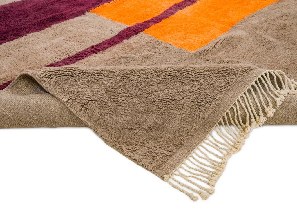 A folded, multicolored rug with fringe detailing on one edge exudes elegance. This luxury rug showcases stripes in orange, purple, and beige on a white background. Its vibrant design adds a touch of sophistication to any room.