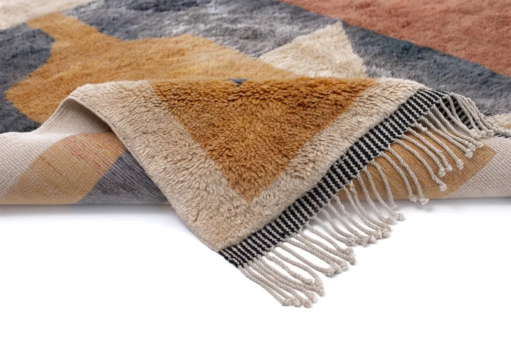 Authentic Moroccan rug with geometric patterns in earthy tones, featuring fringed edges. The hand-knotted piece is partially folded to reveal the exquisite craftsmanship of its underside.