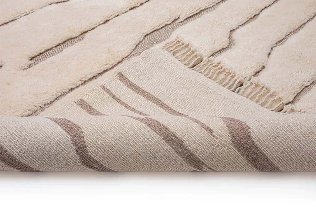 Close-up of an artisanal rug hand-knotted with beige textures and brown stripes, partially rolled to reveal the intricate underside.