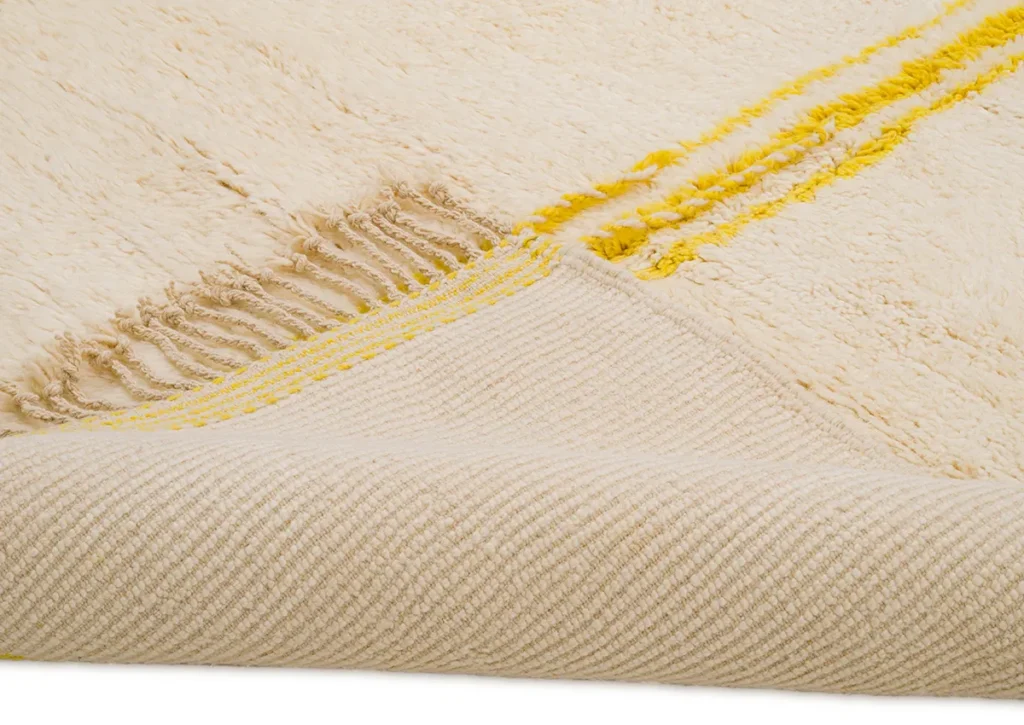 This luxurious, hand-knotted rug features a beige textured design with elegant yellow stripes and fringe on one corner. Partially folded, it reveals the intricate craftsmanship on the underside.