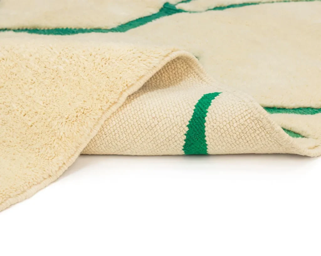 A close-up of a luxurious beige textured rug with a green stripe, partially folded to reveal the reverse. This exquisite piece showcases the intricate craftsmanship typical of hand-knotted rugs.