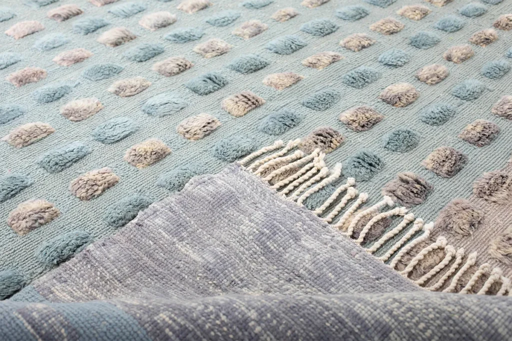 This luxurious hand-knotted rug showcases a close-up of its textured, blue and gray color palette, featuring raised oval patterns and a fringe on one edge.